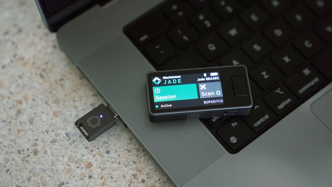 A small Blockstream Jade bitcoin hardware wallet device connected via USB to a laptop keyboard, displaying a "Session" screen.
