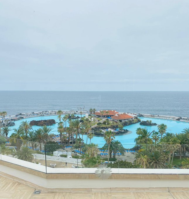 The image shows a beautiful view of an ocean resort with a large pool, palm trees, and other buildings near the water. The sky above appears cloudy, creating a serene atmosphere for relaxation. There are several people enjoying their time at the resort, some closer to the water while others are further away from it. A bird can be seen flying in the distance, adding to the overall charm of the scene.