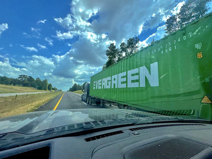 A view from the dashboard of a car driving on a highway, passing a large green Evergreen shipping container marked "EGS U 175740" and "45G1." The image captures the moment of passing the truck, matching the post's caption about overtaking another vehicle on the road.