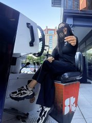 A person wearing black clothing, chunky sneakers, and sunglasses sits on a red and blue cooler outside a white van.