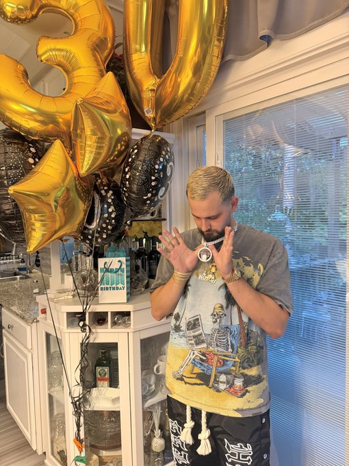 A man with blonde hair and a beard stands in a kitchen looking down at a gift bag, surrounded by gold '30' balloons and a 'Happy 30th Birthday' gift bag.