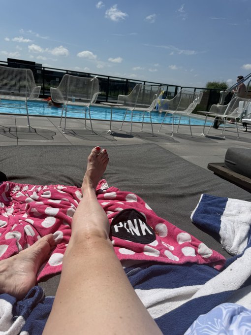 A person's feet are visible resting on a pink polka-dot 'PINK' towel by a swimming pool, with white lounge chairs lined up along the water's edge under a partly cloudy sky.