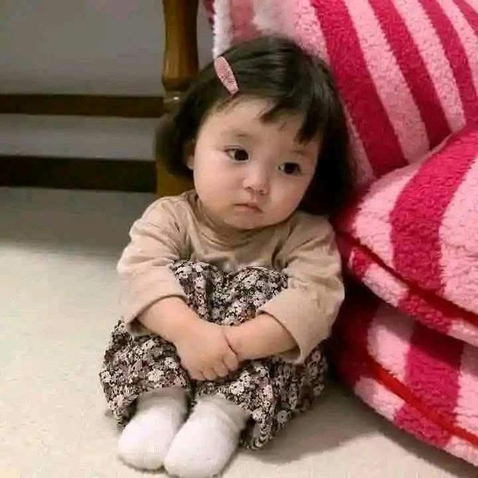 A young child with short dark hair and a pink hair clip sits on the floor leaning against a pink and white striped blanket, wearing a beige top and floral pants with a slightly sad expression.