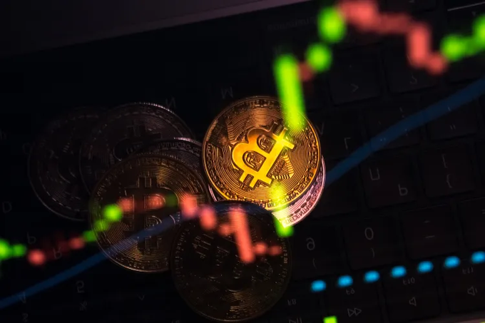 A close-up photograph of physical Bitcoin coins resting on a laptop keyboard, overlaid with a red and green candlestick trading chart.