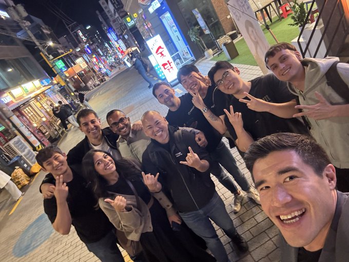 A group of smiling friends and coworkers pose for a selfie on a brightly lit city street at night, many making peace signs and rock-on hand gestures toward the camera.
