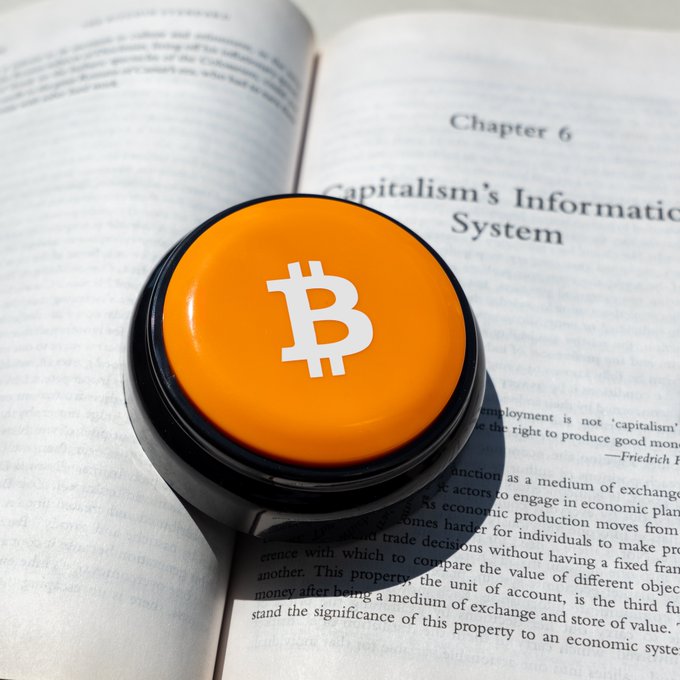 An orange button with the Bitcoin '₿' logo resting on an open book, with the visible page header reading 'Chapter 6 Capitalism's Information System' and a quote attributed to Friedrich H.