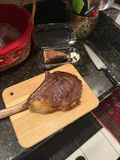 A large tomahawk steak rests on a wooden cutting board next to a knife and what appears to be a chocolate dessert in plastic packaging.