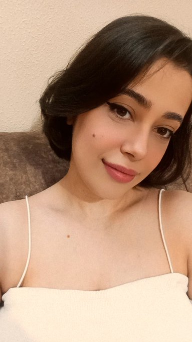 A selfie-style photograph of a young woman with short dark hair, light skin, and subtle makeup, wearing a white top with thin straps against a plain wall.