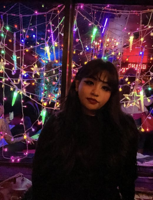 A young woman with long dark hair posing in front of a window decorated with colorful string lights and star ornaments.