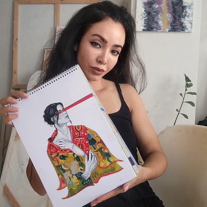 A woman with long dark hair holding up a sketchbook displaying a colorful illustration of a samurai-style figure.