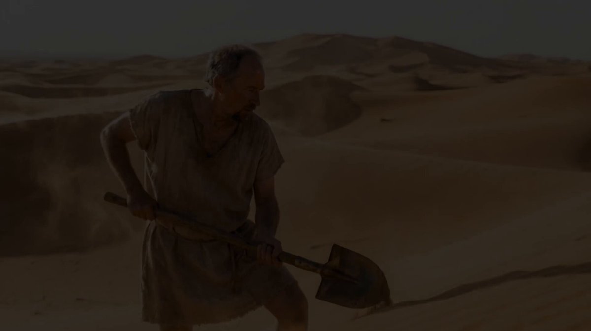 urn of water, a man in a beige robe and headscarf holds a shovel with a handle pointing upwards, set against a desert landscape.