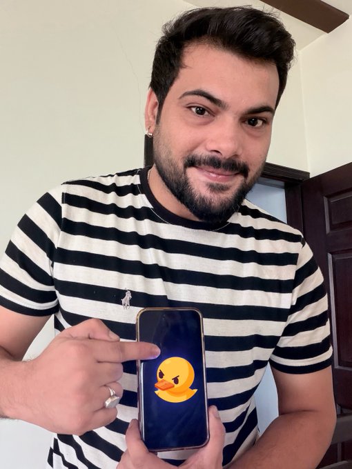 A smiling young man with a beard wearing a black and white striped shirt holds up a smartphone displaying an angry duck emoji on its screen.