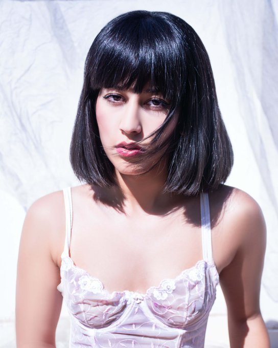 A portrait of a young woman with a black bob haircut and bangs, wearing a lavender camisole top against a white backdrop.