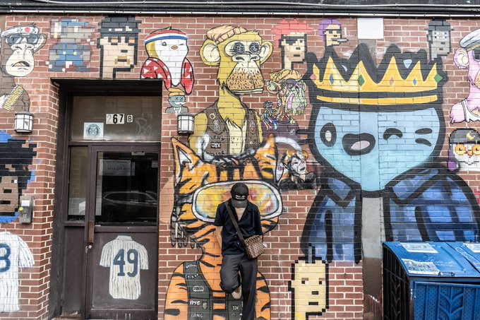 A person wearing a black jacket, cap, and face mask walks past a brick wall covered in colorful street art murals featuring cartoon characters, animals, and a jersey with the number '49'.
