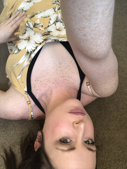 A young woman lies on a carpet wearing a yellow floral top, with visible freckles on her chest and leg. Her body and face are oriented upside-down in the frame.