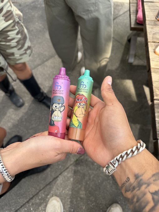 Two hands meet over a sidewalk to hold up colorful "Funboy" branded vape pens featuring NFT monkey character art—one pink and one teal—linking physical product design to the digital NFT collectible theme.