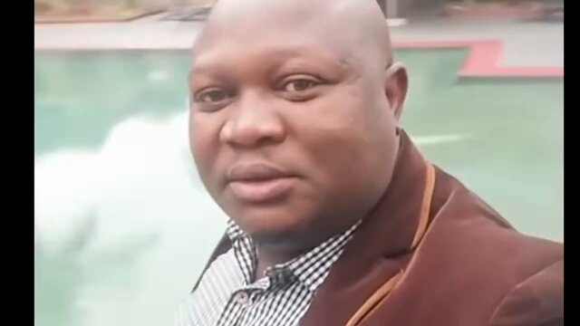 The image features a bald man wearing a brown jacket standing near a swimming pool. The man appears to be looking directly at the camera with an expression of surprise or curiosity. The surrounding area includes a green lawn, suggesting that they are likely outdoors on a sunny day.