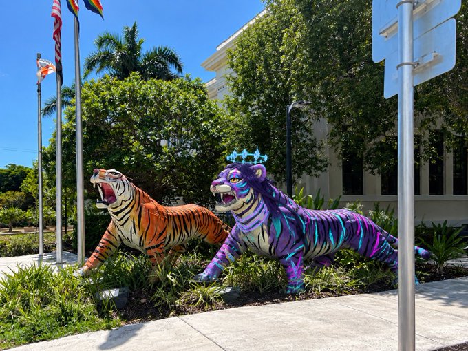 Two colorful, roaring tiger statues—one orange with black stripes and one purple with neon blue stripes wearing a crown—sit in a landscaped garden area in front of a building with flagpoles.