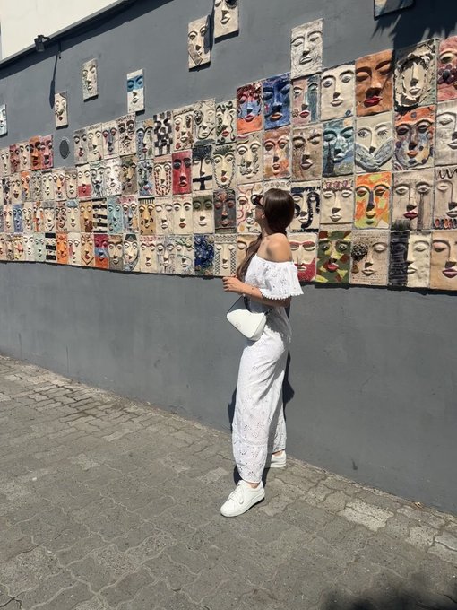 A woman in a white off-the-shoulder top and pants standing on a cobblestone street in front of a wall covered with colorful ceramic face art tiles.