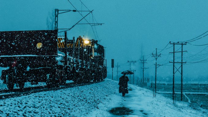 A train with bright headlights moves along snow-covered tracks at dusk while a lone person with an umbrella walks along the adjacent path in falling snow.