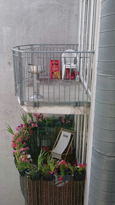 A view from above of a small concrete balcony with a white plastic chair, a birdbath, and scattered red packages, with a lower balcony filled with vibrant pink and purple potted flowers and a wooden lounge chair labeled 'AMA'.