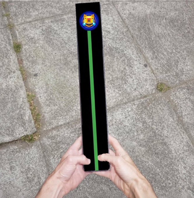 A first-person view of hands holding a long, narrow black rectangular device displaying a Shiba Inu logo at the top and a vertical green line down the center against an outdoor pavement background.