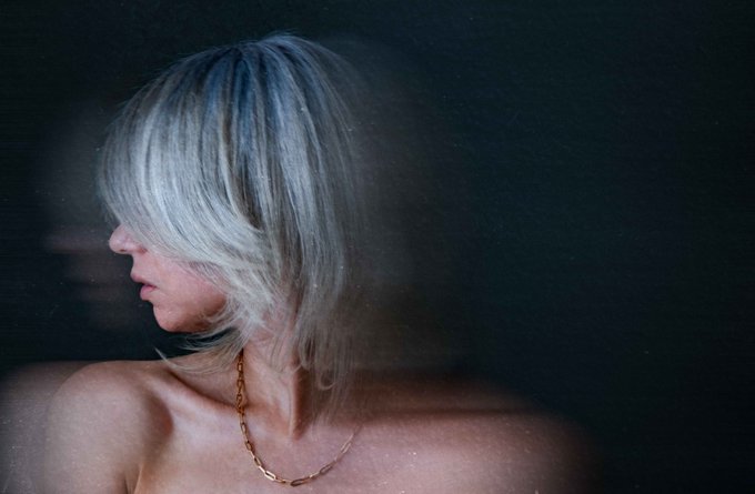 A moody portrait photograph of a woman with short blonde hair in profile, wearing a gold chain necklace, with her face partially obscured by her hair against a dark background.