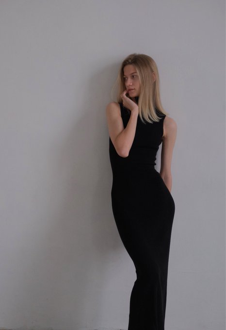 A young woman with long blonde hair stands against a plain grey wall, wearing a sleeveless black floor-length dress, posing with one hand near her chin.