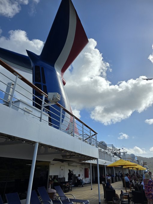 A view from the deck of a cruise ship looking up at a red, white, and blue funnel, with passengers sitting under yellow umbrellas and a plane flying in the cloudy sky.