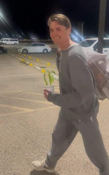 A young man in a gray sweatsuit smiles while walking through a parking lot at night, carrying a small potted plant and a mesh bag, with cars visible in the background.