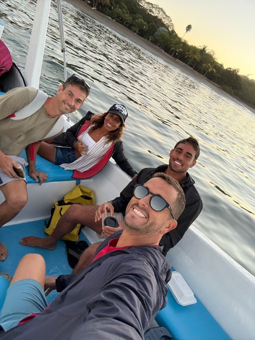 A group selfie of four smiling people on a small boat or watercraft at sunset, with calm water and a tree-lined shore visible in the background.