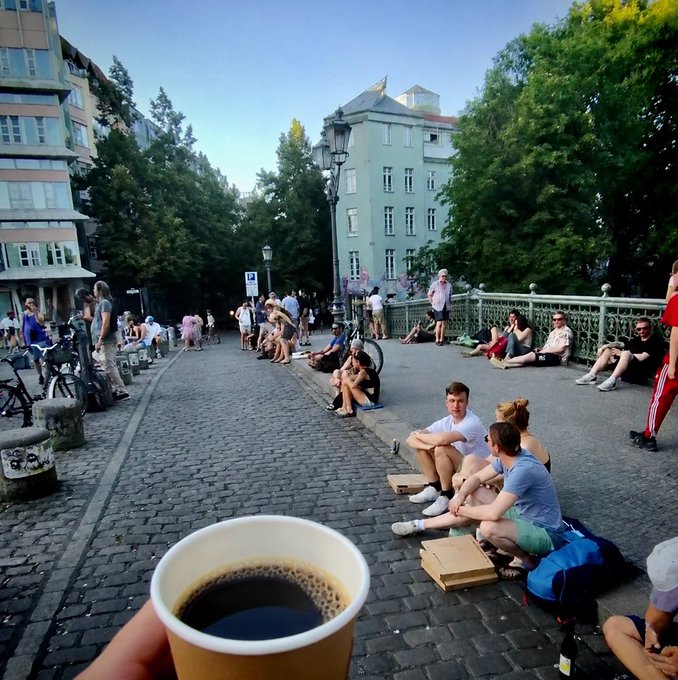 First-person photo of someone holding a paper cup of black coffee in an outdoor European plaza at sunset, with people sitting on the cobblestone bridge railing and green trees in the background.