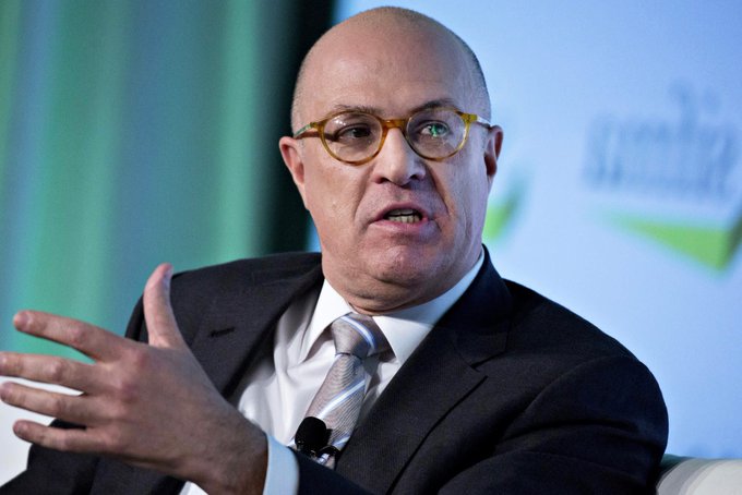 A bald man with yellow-rimmed glasses, wearing a dark suit and striped tie, speaks while gesturing with his hand.