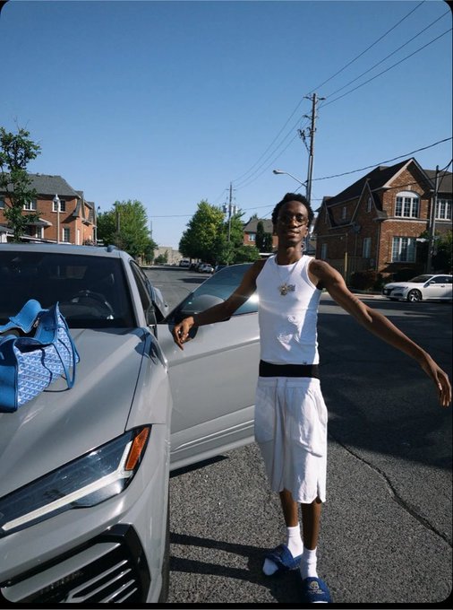 A young man in a white tank top, white shorts, and glasses stands with arms outstretched next to a silver car on a residential street, with a blue bag visible in the car.