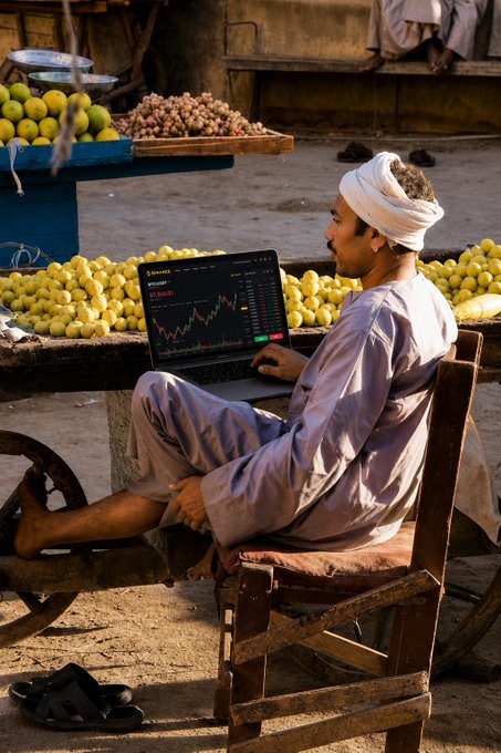 A man in traditional attire sits on a wooden cart at a fruit market, intently watching a Binance trading chart on his laptop showing a price of "$7,208.81." The image captures the post's message perfectly—demonstrating that trading happens anywhere with a laptop and internet connection, even amid crates of citrus fruits.