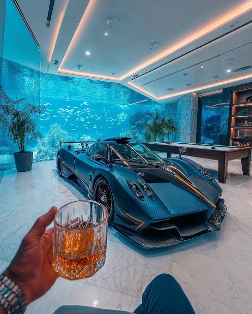 A person holding a glass of whiskey in a luxurious room with a blue supercar parked indoors next to a large aquarium.
