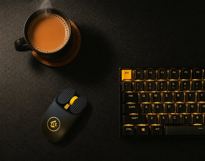 A steaming cup of chai sits on a dark desk beside a black and yellow wireless mouse and a backlit mechanical keyboard featuring an orange "ESC" key, all accented with golden lighting that complements the rich amber color of the tea.
