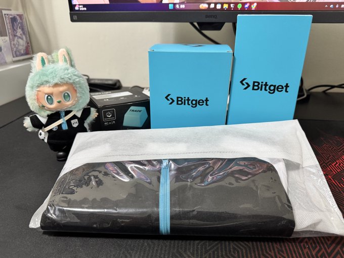 Bitget-branded merchandise boxes and a sealed pouch sit on a desk next to a plush doll, accompanying a post thanking "@yoaka__" for sending Bitget swag after the user was scammed and later filed a police report.