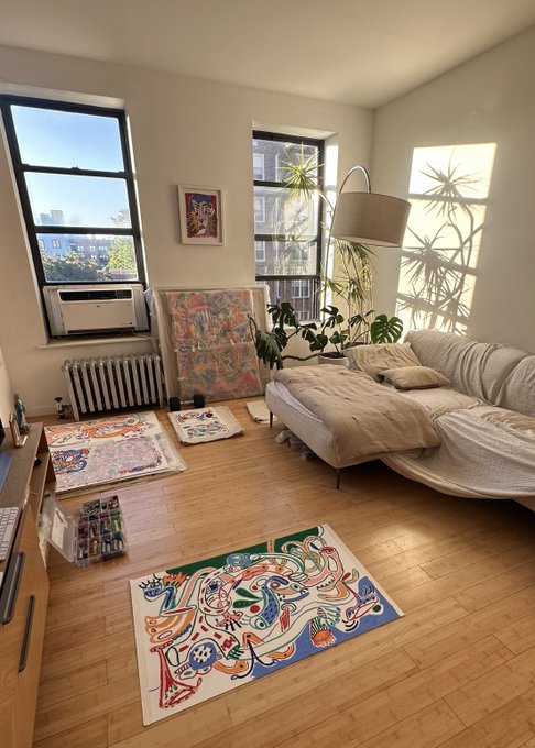 A sunlit, minimalist apartment interior with hardwood floors, featuring a beige sectional sofa, large windows with a city view, a cast iron radiator, and colorful abstract artworks leaning against the walls.