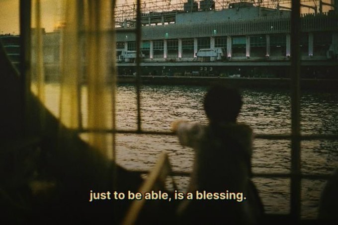 A cinematic shot of a shirtless person viewed through a window, looking out over water toward a stadium at dusk, with the subtitle reading 'just to be able, is a blessing.'