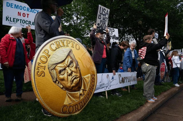 A photo of a protest features several people holding signs, with a large, edited overlay of a gold "Trump" coin in the foreground that says "STOP CRYPTO CORRUPTION" and features Donald Trump's profile. The image illustrates the user's point by using the satirical "Trump coin" as a visual representation of the volatility and speculative nature of memecoins.