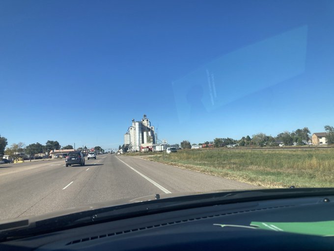 View from a car windshield driving on a multi-lane highway toward a grain elevator complex under a clear blue sky.
