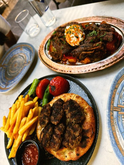 A plate of grilled meat kebabs served on flatbread with french fries, roasted tomatoes, and green peppers, alongside a larger platter of grilled meats topped with a creamy sauce.