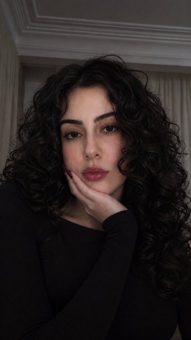 A portrait of a woman with long, curly dark hair posing with her hand against her cheek, wearing a black top against a softly lit background with curtains.