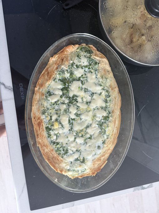 A freshly baked spinach and cheese pie or quiche in a glass oval dish, sitting on a black stovetop next to a pan of cooking food.