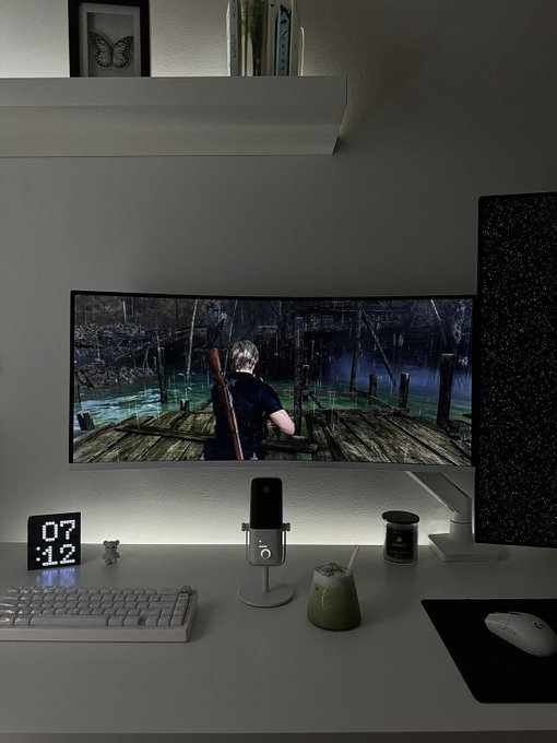 A gamer's desk setup features an ultrawide monitor displaying Resident Evil 4's Leon on a dock, accompanied by a keyboard, mouse, microphone, green tumbler, and a flip clock reading "07" — perfectly capturing the early morning "Good morning homies, happy Friday" vibe as they gear up for a fruitful day of gaming.