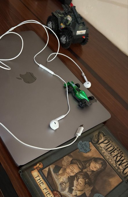 A lifestyle photo of a silver MacBook, white wired earphones, a small green toy race car, a toy tow truck, and a Spider-Man book arranged on a dark wooden surface.