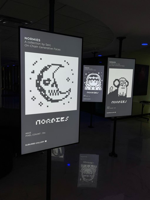 An art gallery displaying illuminated screens featuring pixelated skull artwork titled 'NORMIES - A collection by Serc On-Chain Generative Faces' with details like '#605 PIXEL COUNT: 394'.