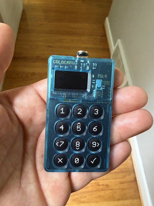 A hand holds a translucent blue COLDCARD Mk4 hardware wallet with a numeric keypad, including 1–9, directional arrows, ×, 0, and ✓. The joke references Schrödinger’s cat: the wallet’s private keys may feel simultaneously “yours and not yours,” since possessing the device does not prove the keys inside are usable.