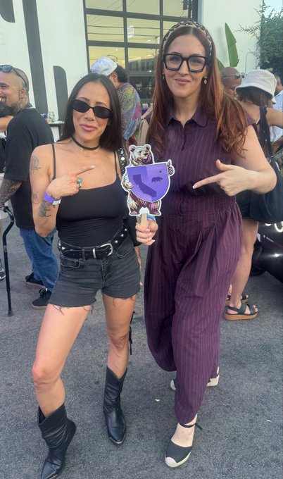 Two women posing at an outdoor event; one in a black tank top and shorts pointing to a purple tiger sticker held by her companion in a striped purple jumpsuit.